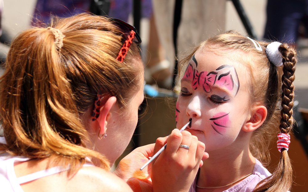Face painter painting a pink butterfly design on a smiling girl at an event