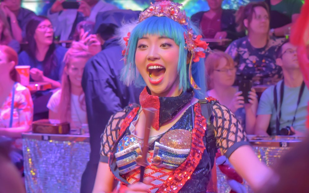 Performer with bright blue hair and a sequined costume on a stage under colored lights